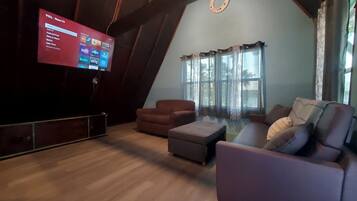 A living room with a TV displaying a streaming service interface, a brown sofa, a grey ottoman, and a window with curtains.