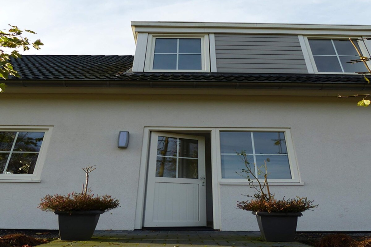 A two-story house with a white exterior, a dark roof, and a front door. There are two potted plants on either side of the door.