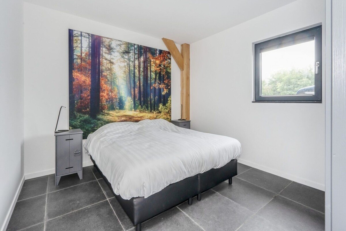 A bedroom with a bed, a nightstand, a large forest-themed painting, a wooden beam, and a window.