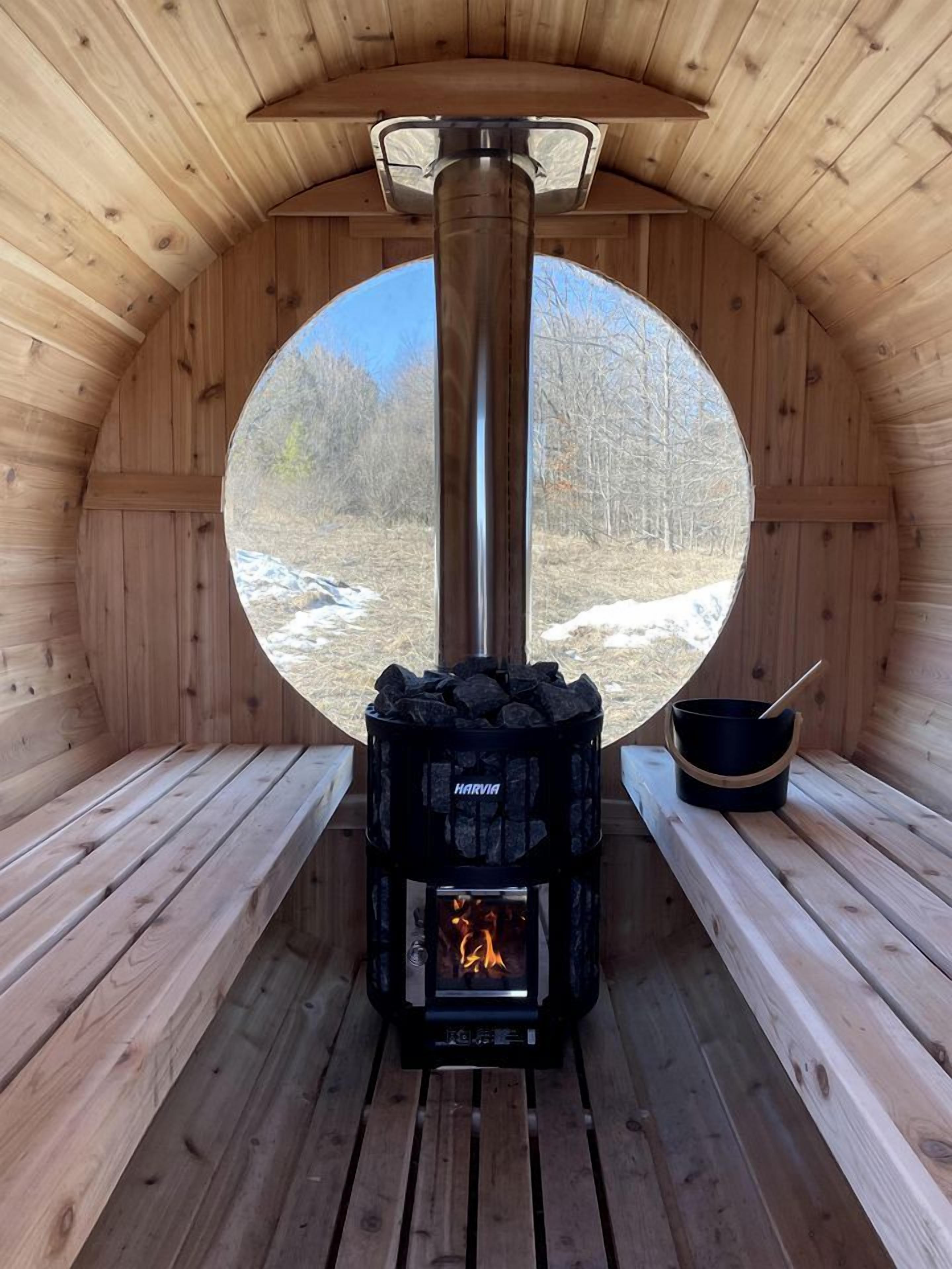 A wooden sauna with a Harvia wood stove, benches, and a view of a snowy landscape.