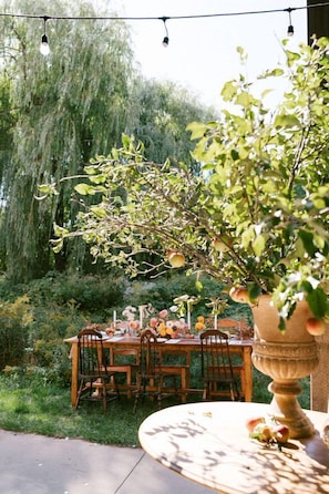 An outdoor dining area with a table set for four, surrounded by chairs, in a garden with a large tree and a decorative urn. - A-Frame Serenity: Tiny Cabin with Panoramic Scenery in Michigan's Tranquil Setting (Boyne Falls)