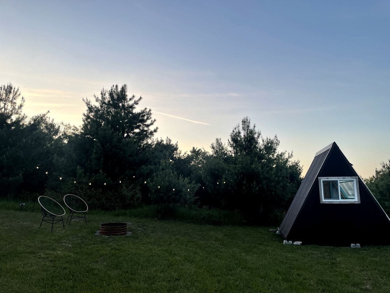 A triangular tent-like structure in a grassy area with two chairs and string lights.