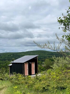 A small, dark-colored cabin with a sloped roof, two doors, and a chimney, surrounded by greenery and hills. - A-Frame Serenity: Tiny Cabin with Panoramic Scenery in Michigan's Tranquil Setting (Boyne Falls)