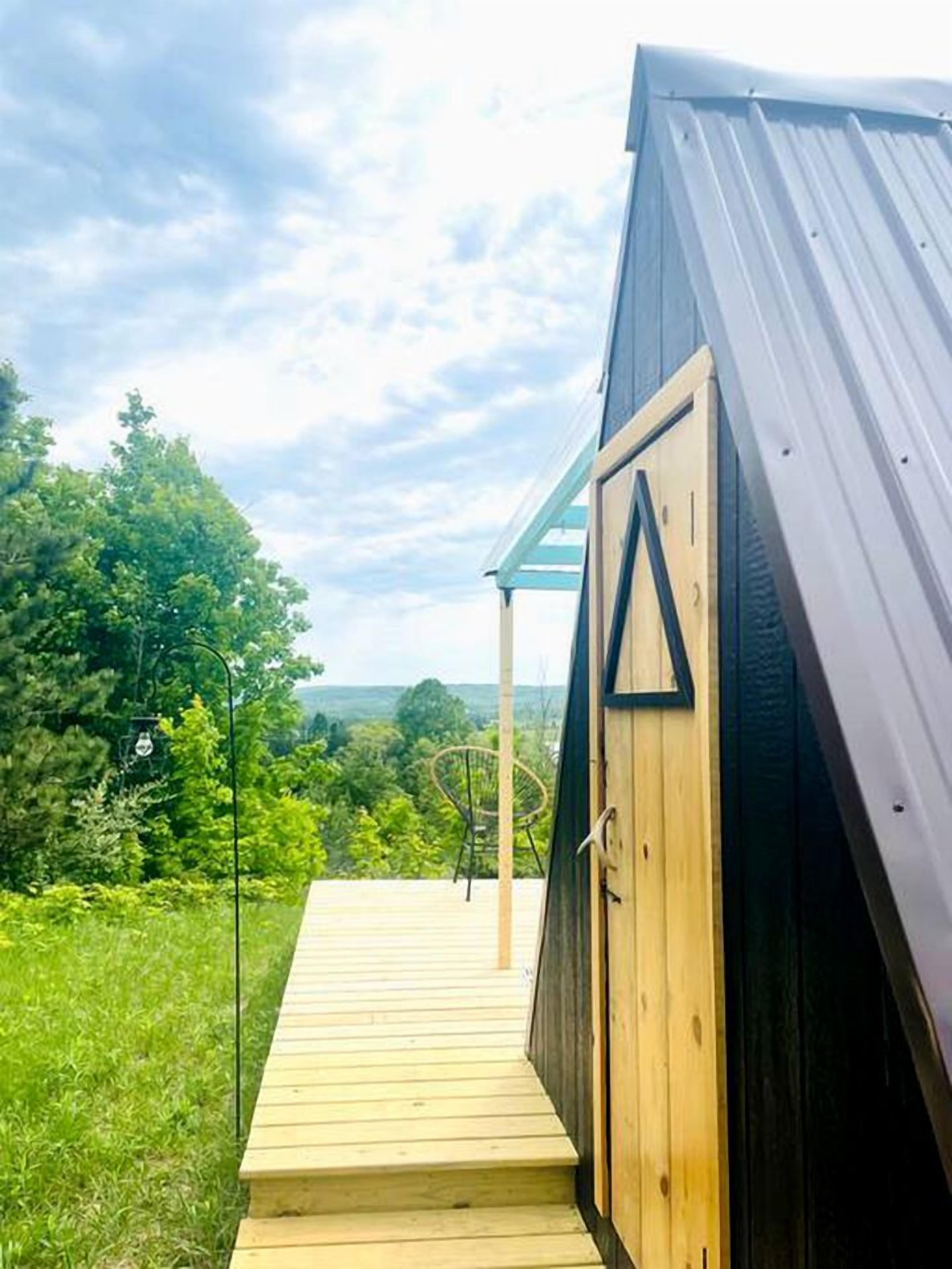 Una terraza de madera que conduce a un edificio de color oscuro con una puerta triangular, rodeado de follaje y un cielo despejado.