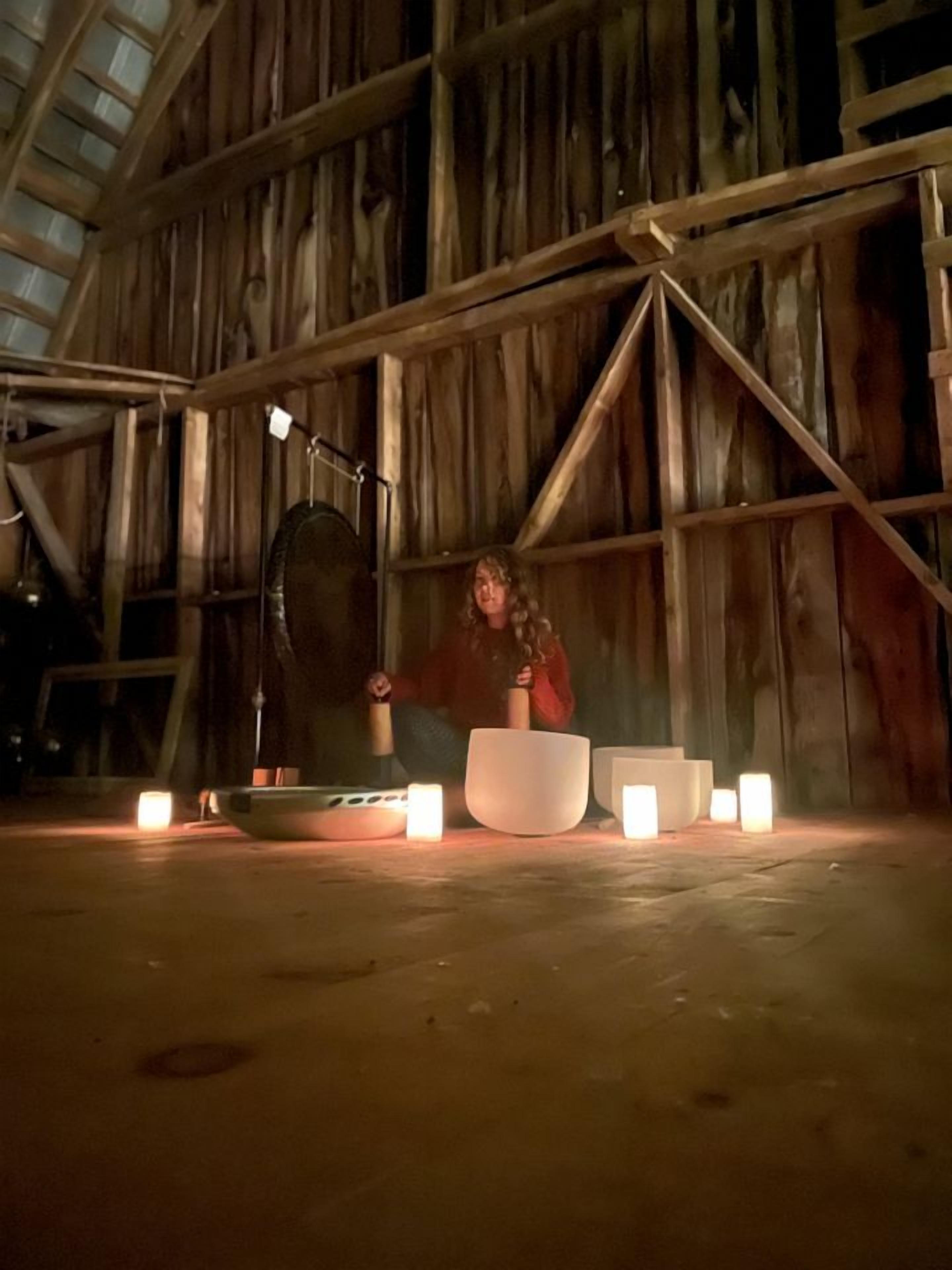 A rustic interior with wooden beams, a large white bowl, and several lit candles on a wooden floor.