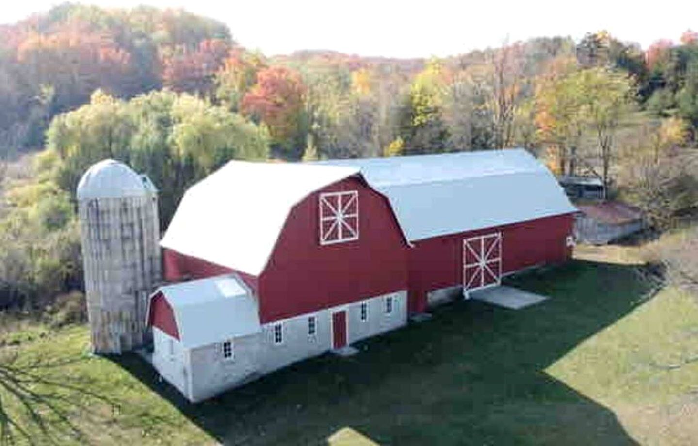 A red barn with white trim and a silo, surrounded by a grassy field and trees.