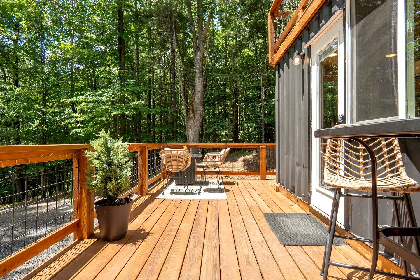 Una terraza de madera con mobiliario exterior, una planta en maceta y vistas a un área boscosa.