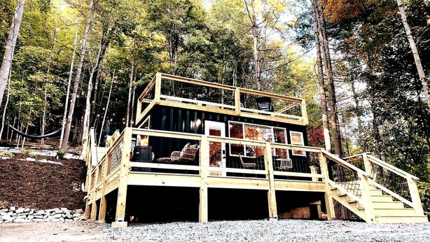 Una cabaña de madera de varios niveles, con balcón y escaleras, rodeada de árboles.