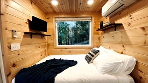Un dormitorio de cabaña de madera con cama, ventana, aire acondicionado y televisor montado.