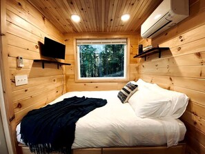 A wooden cabin bedroom with a bed, a window, an air conditioner, and a mounted TV. - Tiny Luxe Living: Todd, NC Retreat with Hot Tub and Breathtaking Deck Views (Todd)