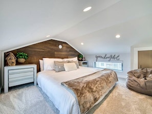Ein Schlafzimmer mit schrägverdachter Decke, einem Bett mit weißer und brauner Decke, einem Nachttisch mit Lampe und einer Wand mit der Aufschrift 'goodnight'.