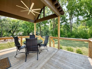 Eine Holzterrasse mit Tisch und Stühlen, umgeben von einem Hollerdgeschoss, mit Blick auf eine grüne Wiese mit Bäumen.