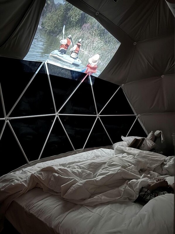 A tent with a geometric window design, a bed with white bedding, and a view of a river with boats.