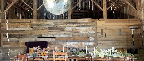 L’intérieur d’une grange rustique présente une table à manger en bois, dressée pour six personnes, entourée de chaises en bois. Une grande boule à facettes est suspendue au plafond, et les murs sont recouverts de planches de bois.