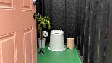 Um corredor estreito com piso verde, uma porta fechada, um vaso de planta e uma lixeira.