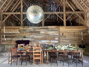A rustic barn interior with a wooden dining table set for six, surrounded by wooden chairs. A large disco ball hangs from the ceiling, and the walls are made of wooden planks.