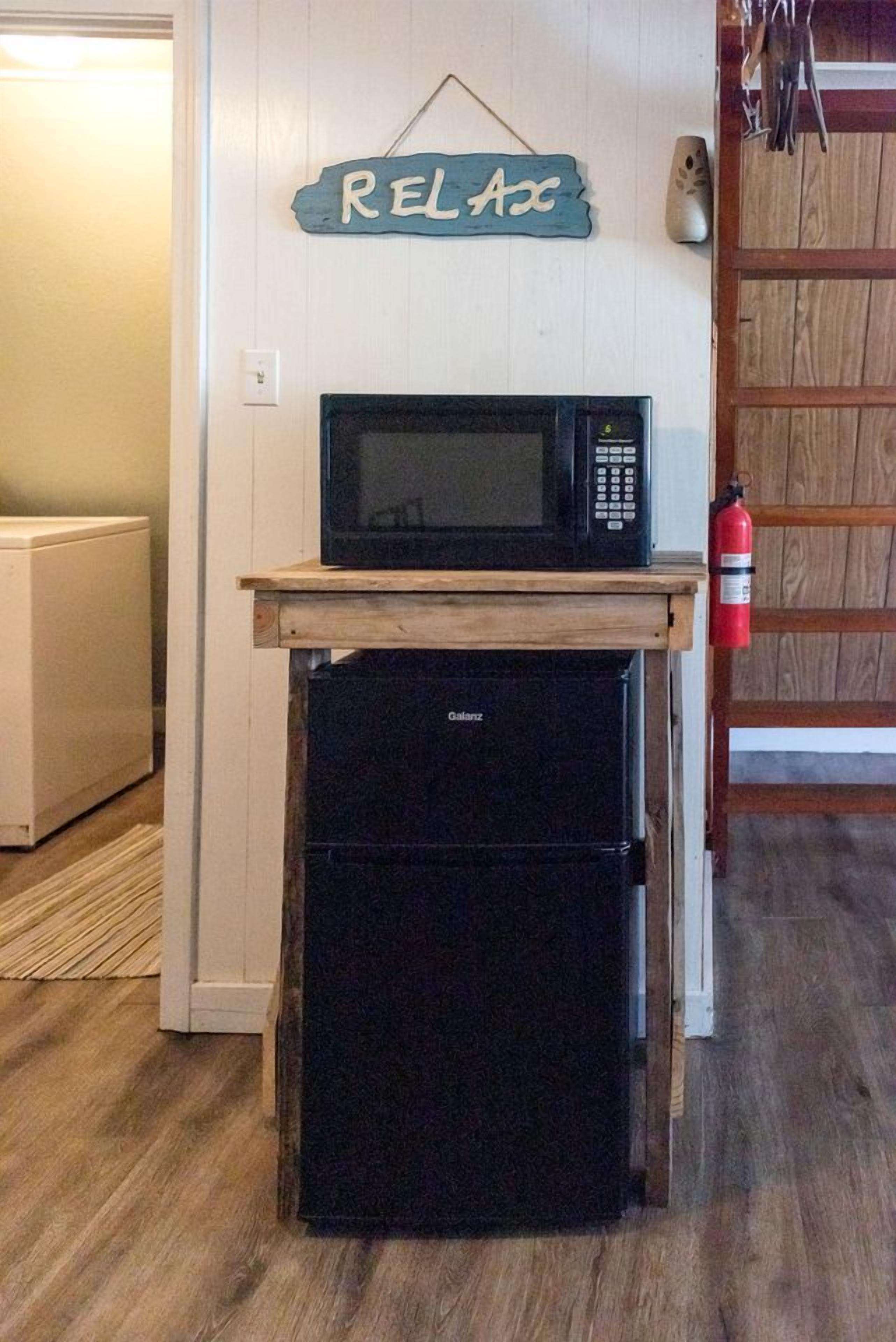 A kitchen with a microwave and a refrigerator on a wooden countertop, a 'RELAX' sign on the wall, and a fire extinguisher mounted on the wall.