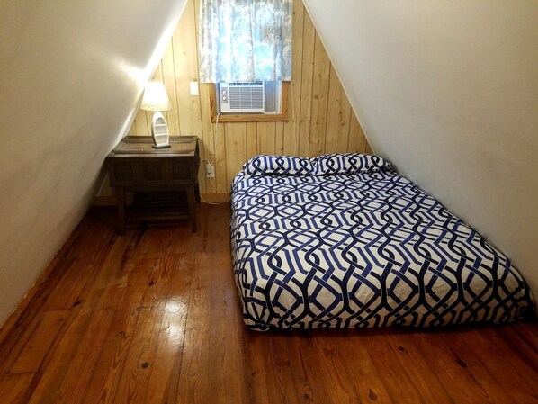 A small, sloped-roofed room with a wooden floor, a bed with a geometric-patterned bedspread, a bedside table with a lamp, and a window with a curtain.