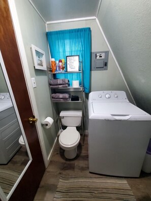 Une petite salle de bain comprenant des toilettes, un lave-linge et une étagère avec des articles de toilette.