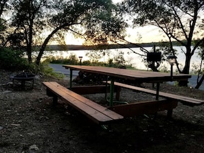A picnic table with benches, a fire pit, and a lamp post near a body of water.