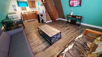 A compact living space with a kitchen, a dining area, a sofa, a wooden table, and a TV mounted on the wall.