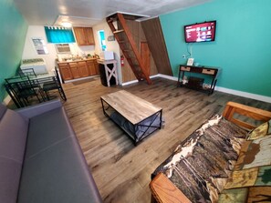 A compact living space with a kitchen, a dining area, a sofa, a wooden table, and a TV mounted on the wall.