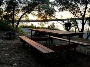 Una mesa de picnic con bancos, un hogar para fogata y un farol cerca del agua.
