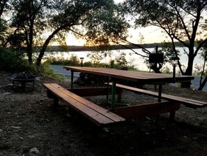 Una mesa de picnic con bancos, un hogar para fogata y un farol cerca del agua.