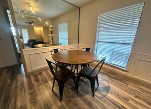 A dining area with a round table and four chairs, a kitchen in the background, and a large window with blinds. - *Extended Stay: Backyard, Garage, Fireplace* (Carrollton)