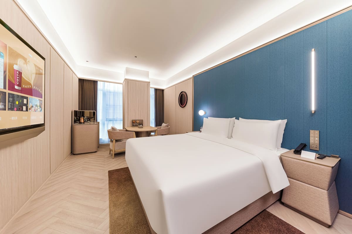 Een hotelkamer met een groot bed, nachtkastje, bureau met stoel, een klein keukenhoekje en een groot raam.