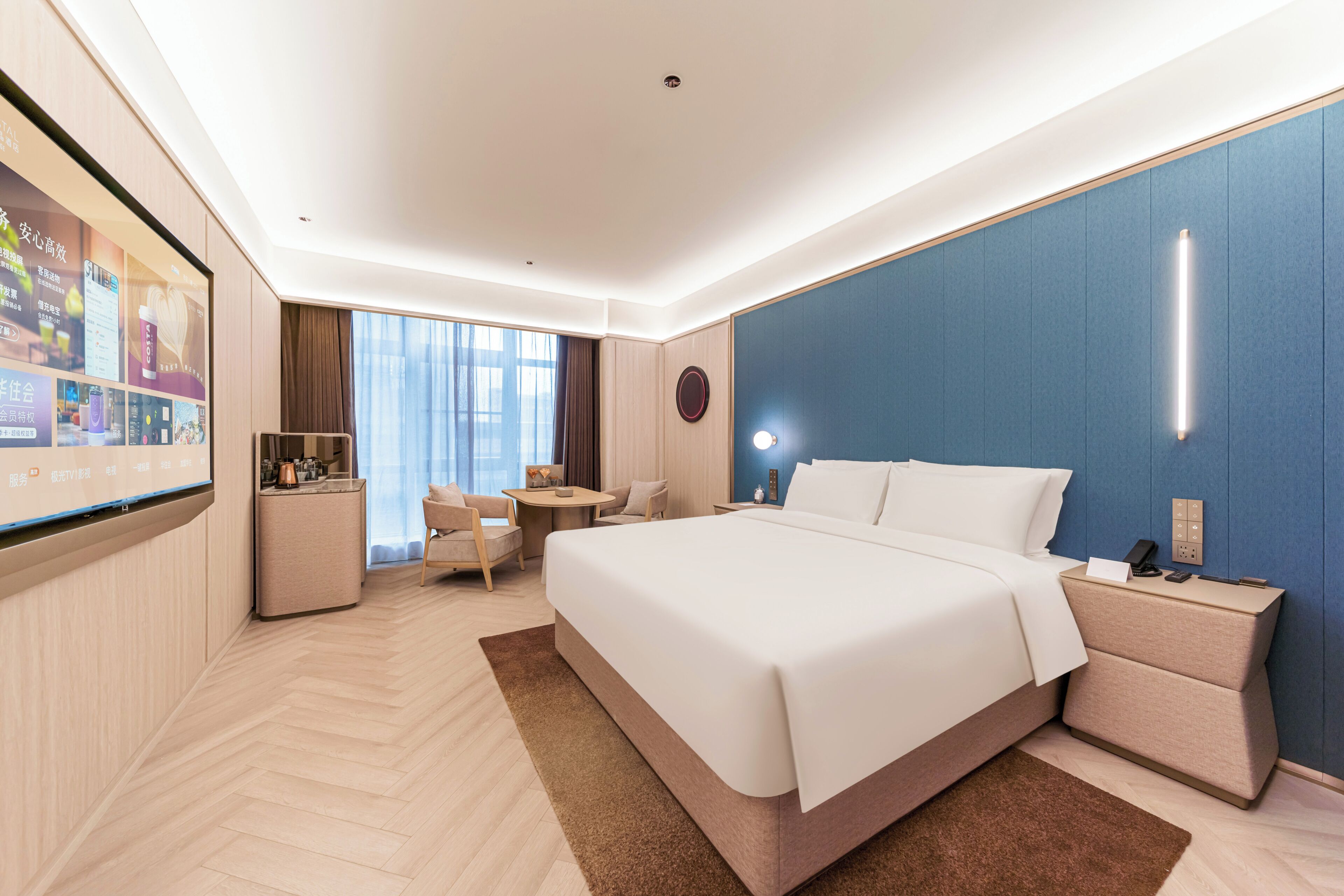 A modern hotel room with a large bed, a desk, a chair, and a TV screen displaying content.