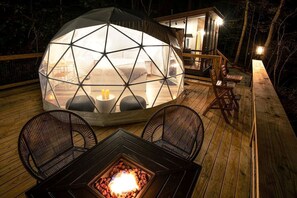 Une structure en forme de dôme géodésique, dotée d’un foyer et d’un espace salon aménagé sur une terrasse en bois.