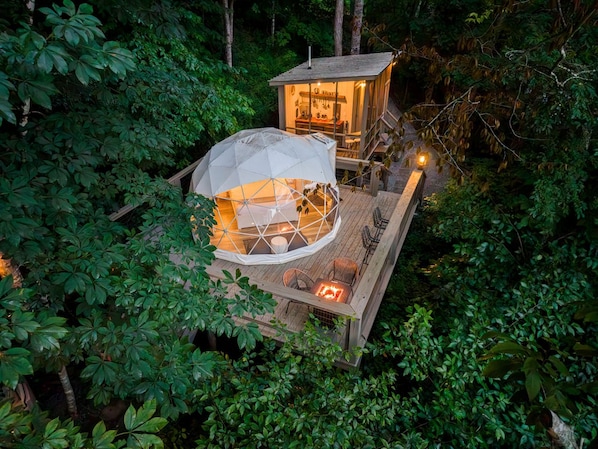 Estructura de domo geodésico con una terraza de madera y zona de estar, rodeada de follaje denso.