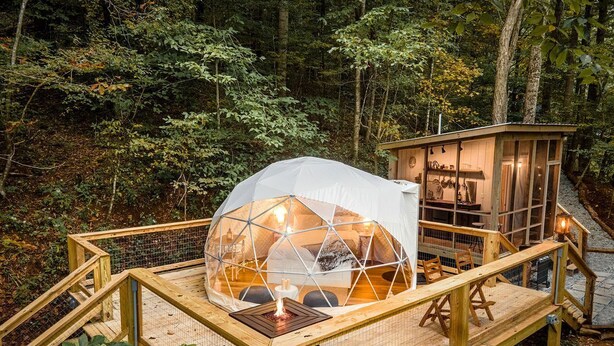 A geodesic dome structure with a wooden deck and a small building with a lighted interior.