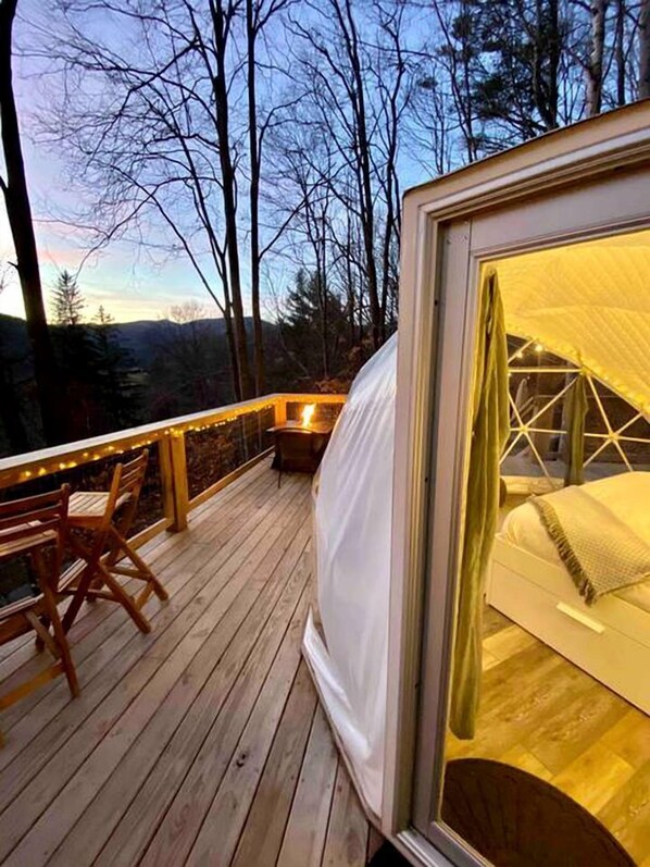 A cozy outdoor deck with a bed, wooden chairs, and a view of trees and hills.