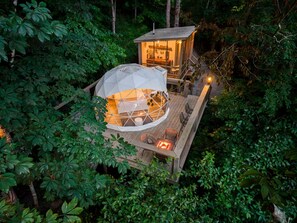 A geodesic dome structure with a wooden deck and seating area, surrounded by dense foliage.