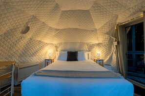 Una habitación con forma de tienda de campaña, una cama, dos mesitas de noche con lámparas y un ventanal.