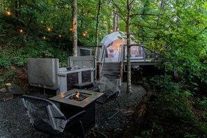 An outdoor seating area with a fire pit, chairs, and a covered structure in a forest setting.