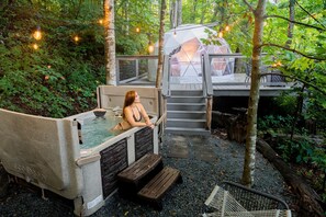 A hot tub in a forested area with a transparent dome structure and a wooden bench.