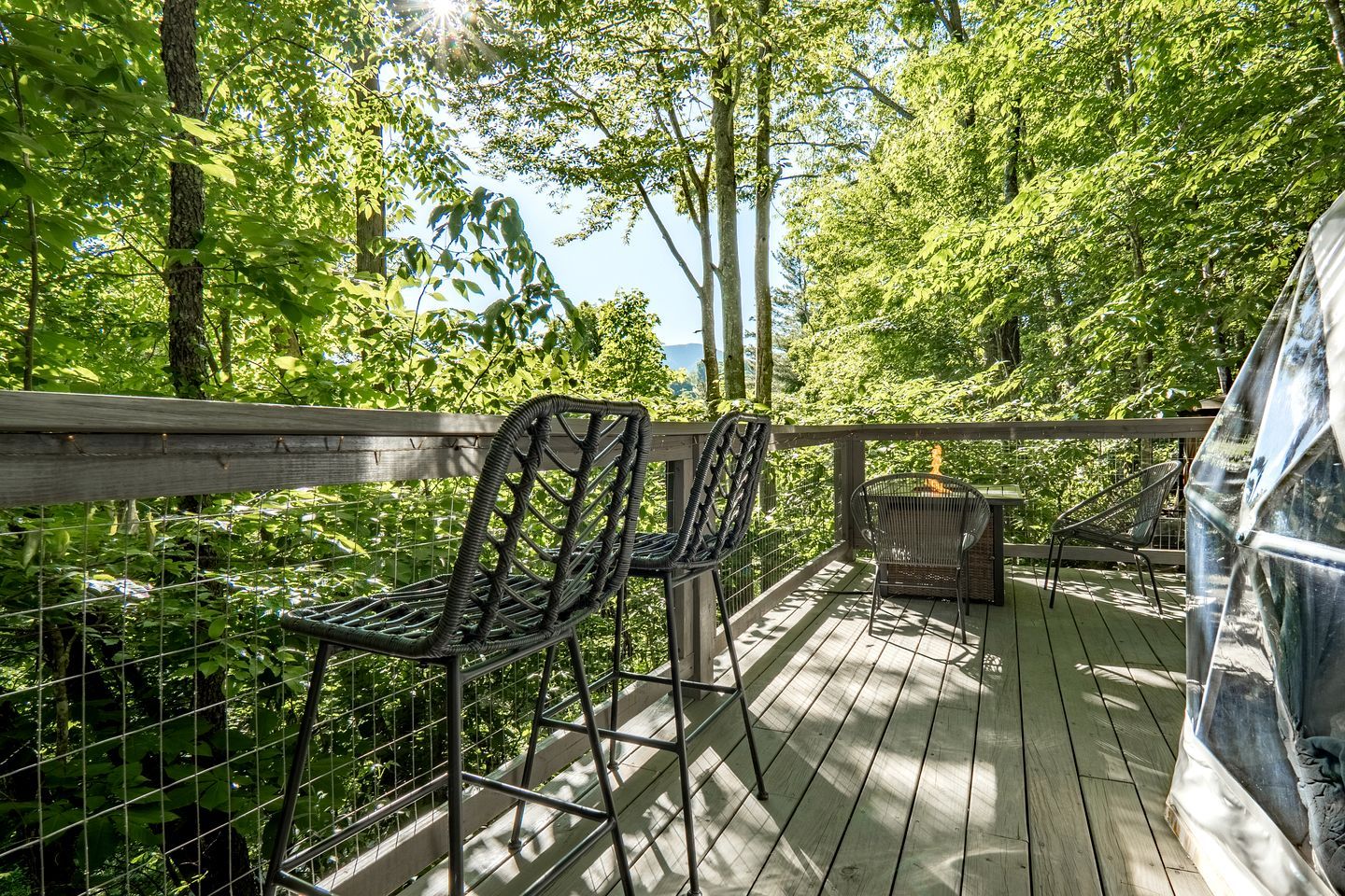 Une terrasse en bois, dotée de chaises et d’un barbecue, entourée d’arbres et offrant une vue sur le ciel.