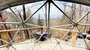 A geodesic dome structure with a wooden deck, outdoor seating, and a view of the surrounding forest.