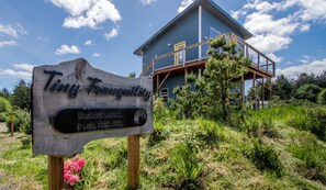 A wooden sign with 'Tiny Tranquility' and 'Vacation Rentals & Long Term Rentals' text, a blue house with a wooden deck, and a clear blue sky.