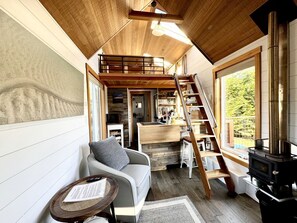 A cozy cabin interior with a sofa, a small table, a wooden staircase, and a wood-burning stove.