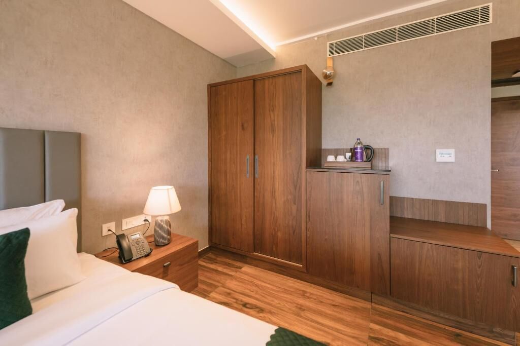 A hotel room with a bed, a nightstand with a lamp, a telephone, a wardrobe, and a small table with a kettle.