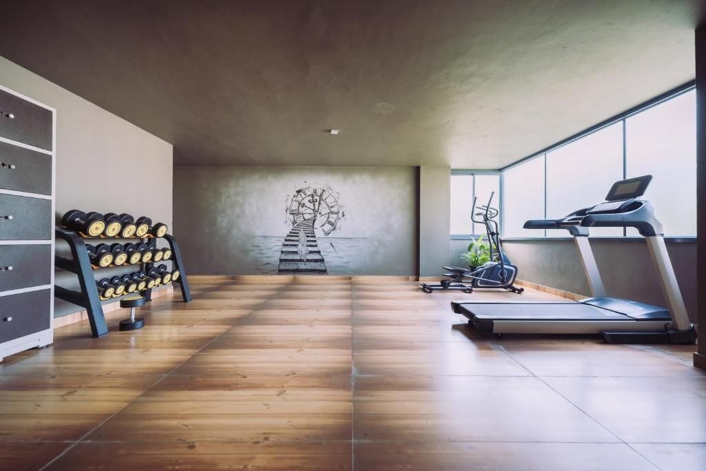Un gimnasio moderno con suelo de madera, un soporte de mancuernas en la pared, una cinta de correr y un ventanal grande.