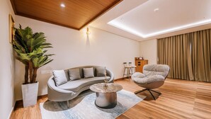A modern living room with a curved sofa, a round coffee table, a patterned armchair, a large potted plant, and a view of a balcony with curtains.