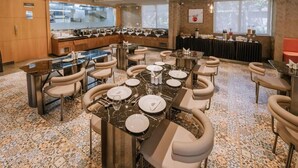 A spacious dining area with multiple tables and chairs, a kitchen in the background, and large windows providing natural light.