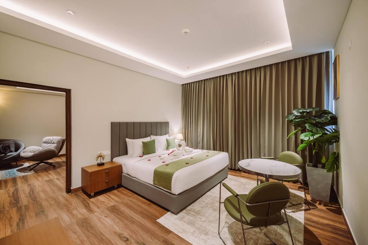 A modern hotel room with a large bed, a round table, a chair, a small side table, and a potted plant.
