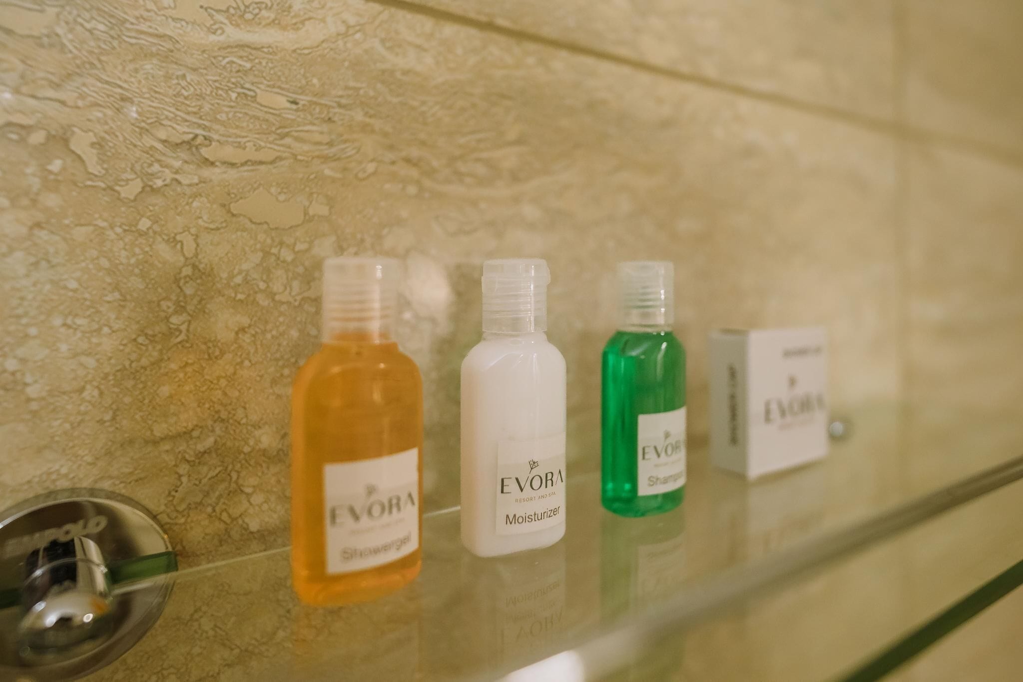 Et badeværelse med en glashylde, der indeholder fire flasker Evora produkter: shower gel, moisturizer og shampoo.