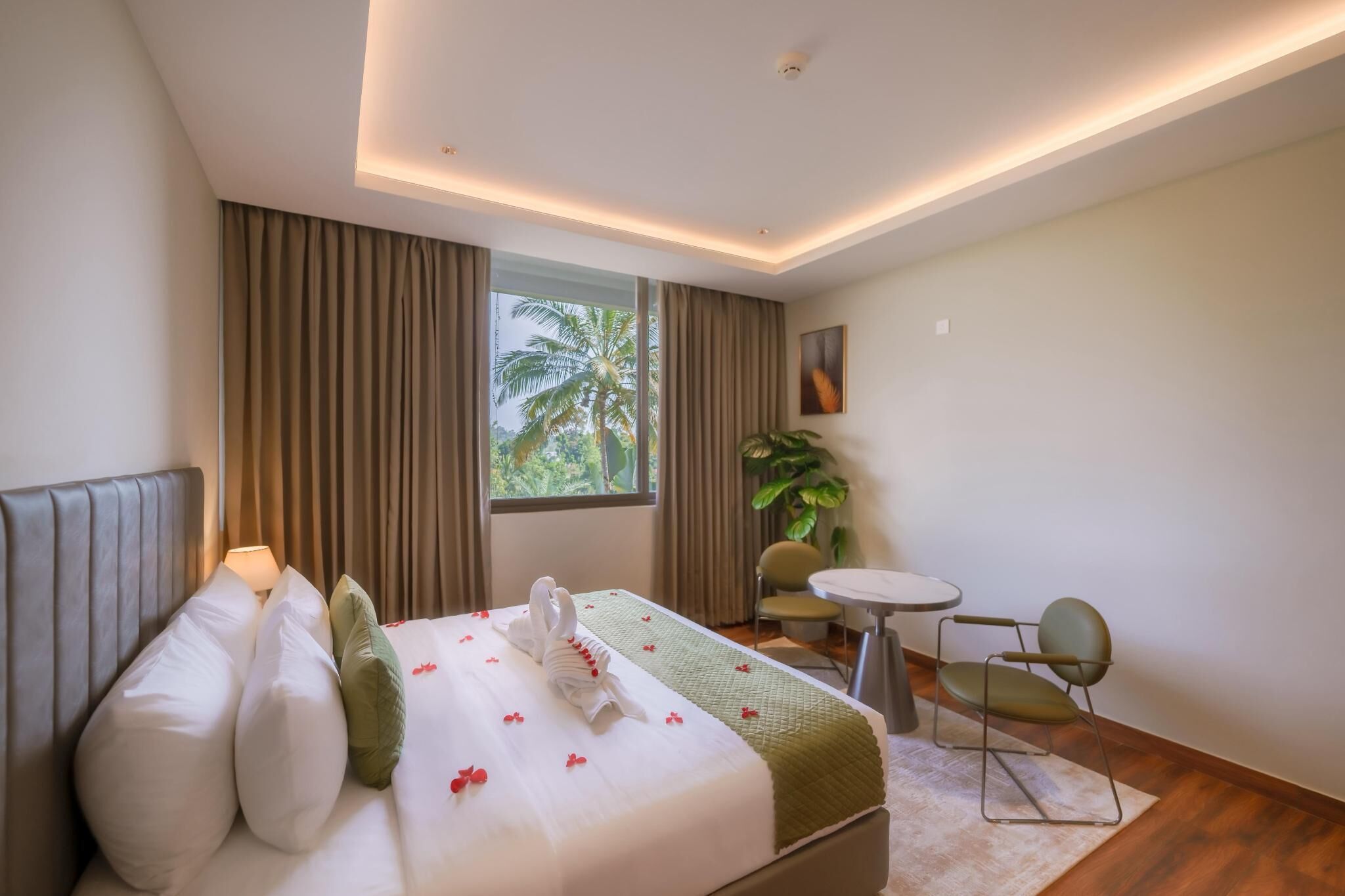 A hotel room with a bed, a small round table, a chair, a potted plant, and a window with a view of palm trees.